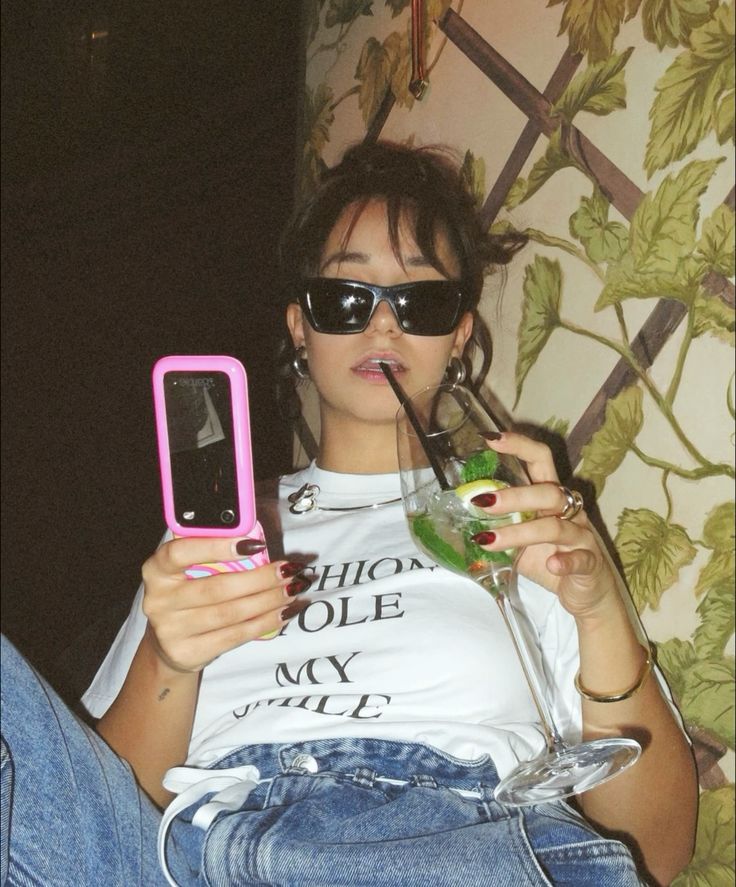 Femme stylée en lunettes de soleil, tenant un cocktail et un téléphone à clapet rose, ambiance détendue
