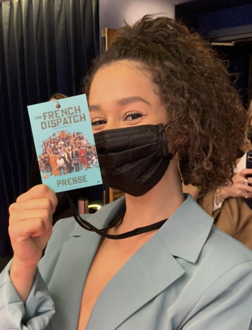 Femme souriante portant un masque noir et un blazer bleu, tenant un badge presse ‘The French Dispatch’.